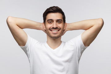 Young man standing isolated on gray background with arms behind head, relaxing peacefully in free time