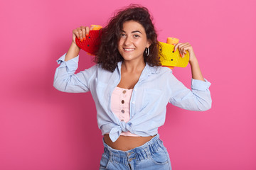 Close up of brunette charming woman with tender smile, holding skateboard on her shoulders, wearing t shirt, shirt and jeans, enjoys hobby, isolated over pink background, ready to skate in park.