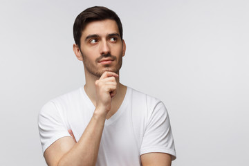 Young guy isolated on grey background standing in blank T-shirt against wall, pressing fist to chin as if dreaming of something, looking pensive and happy