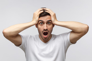 Emotional man with strong expression of fear, round eyes, open mouth and hands pressed to top of head, showing astonishment and shock, isolated on gray background