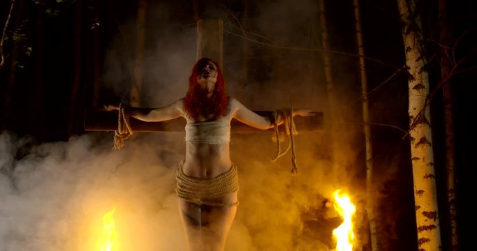 battered young woman with red hair who was crucified on the cross in the woods at night under the light of fire