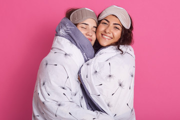 Indoor shot of smiling women with sleep masks, wearing blanket stands hagging isolated over rose...