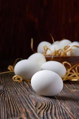 White eggs on a wooden table. In the background are eggs in a wooden bowl and on paper straw. Close up. Low key.