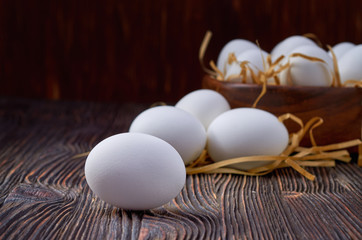 White eggs on a wooden table. In the background are eggs in a wooden bowl and on paper straw. Close up. Low key.