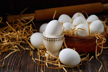 White eggs in a wooden bowl, on a paper straw and a wooden table. On the back of a rolling pin. Low key.
