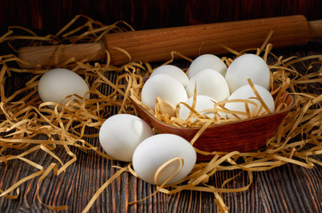 White eggs in a wooden bowl, on a paper straw and a wooden table. On the back of a rolling pin. Low key.