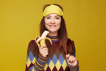Studio portrait of happy young female holding fresh banana in hand, wearing sleeping mask on head after having good nap, has positive face expression, feeling new energy and ready for new experience.