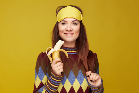 Studio portrait of happy young female holding fresh banana in hand, wearing sleeping mask on head after having good nap, has positive face expression, feeling new energy and ready for new experience.