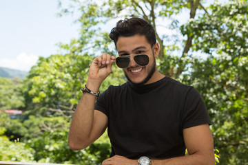 Young latin man portrait indoors next to a window with nature as background. He is smiling He is taking his sunglasses off.. 