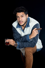 Young latin man studio portrait. The man is standing with one knee up. He has his jacket open. He is holding a glass of scotch and a cigar.
