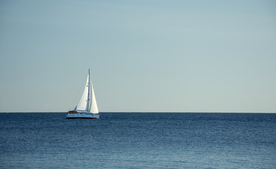 Fototapeta premium Summer day on the Adriatic sea. In the distance, a white sailboat is sailing along the sea in the windy and sunny day. Horizontal, minimalism photo. In shades with blue. With a lot of space for text.