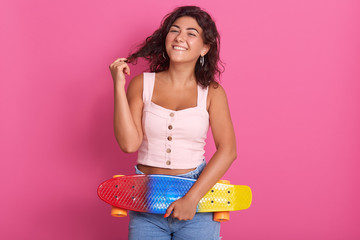 Indoor studio shot of sincere charming model posing over pink background, touching her hair, standing with her skateboard, wearing pink top and jeans, looking directly at camera. Youth concept.