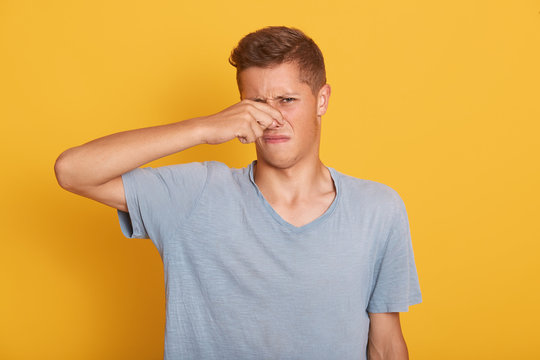 Portrait of young handsome boy having unpleasant facial expression, looking directly at camera, closing his nose with fingers, avoiding awful smell, standing isolated over yellow background in studio.
