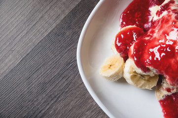 Closeup Vanilla ice cream poured with strawberry or raspberry jam with sliced banana slices on a white plate on a dark wooden table. You can do this at home on vacation