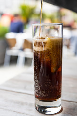 Pouring cola in a long drink glass with, slice of lemon and ice cubes. 