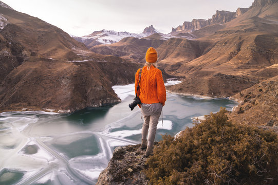 Portrait from the back of the girl traveler photographer in an orange sweater and hat with a camera in hand in the mountains against the background of a frozen mountain lake. Photo travel concept