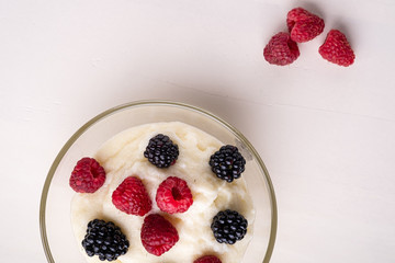 Semolina pudding porridge dessert with  blackberry and raspberry in glass bowl near with scattered heap berries on white wooden background, breakfast, isolated, top view