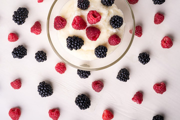 Semolina pudding porridge dessert with  blackberry and raspberry in glass bowl near with scattered heap in order berries on white wooden background, breakfast, isolated, top view