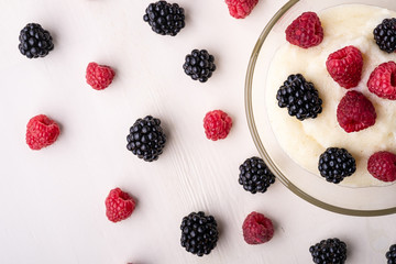 Semolina pudding porridge dessert with  blackberry and raspberry in glass bowl near with scattered heap in order berries on white wooden background, breakfast, isolated, top view