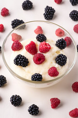 Semolina pudding porridge dessert with  blackberry and raspberry in glass bowl near with scattered heap in order berries on white wooden background, breakfast, isolated, selective focus