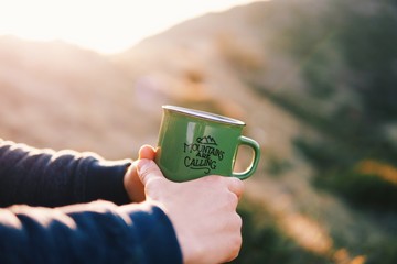 Close up shot of the tourist hands holding green mug with hot drink, traveler holds in hands a mug...