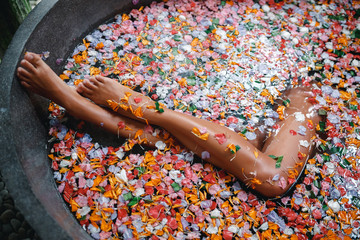 Female legs in bathtub with flower petals and beauty products on wooden tray. Organic spa relaxation in luxury Bali outdoor bath