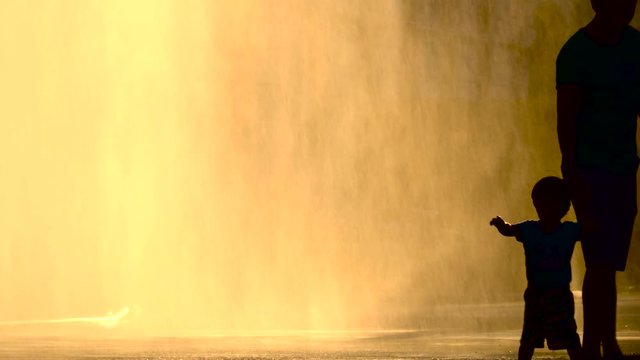 Father and son silhouetted against the late evening sun as they cool off by a fountain in the middle of a heatwave, static shot