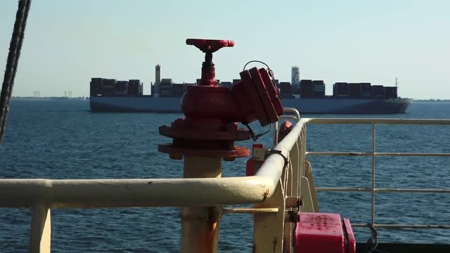 RIGA/LATVIA - JUNE 21 2019: Red valve above thin yellow rusty pipes against long container ship sailing among ocean with horizon on background on June 21 in Riga