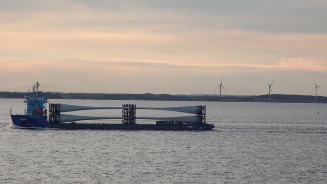 RIGA/LATVIA - JUNE 21 2019: Long hopper barge sails on sea surface against land covered with high windmills row under pink sky at sunset on June 21 in Riga