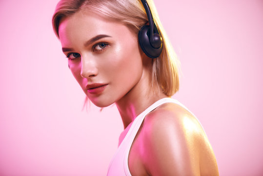 Music in my soul. Close up of cute blonde woman in headphones listening music and looking at camera while standing against pink background