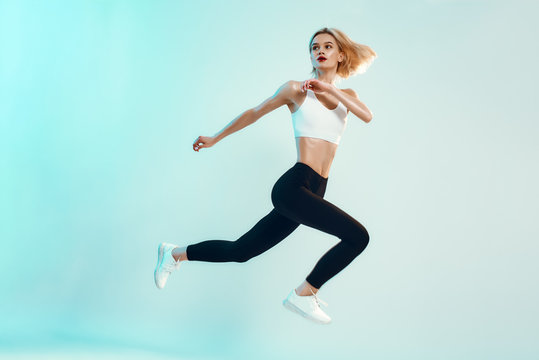 No limits! Sporty and young woman with perfect body in white top and black leggings jumping against blue background in studio