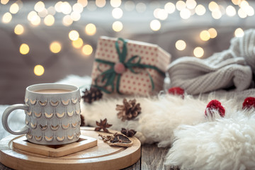 Coffee cup over Christmas lights bokeh in home on wooden table with sweater on a background and present. Holiday decoration, magic Christmas