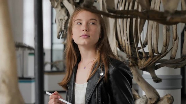 Teenager cute girl makes draw sketches in graphic tablet of animal skeletons in museum of nature as part of school task of biology