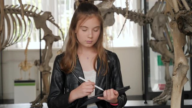 Teenager cute girl makes draw sketches in graphic tablet of animal skeletons in museum of nature as part of school task of biology