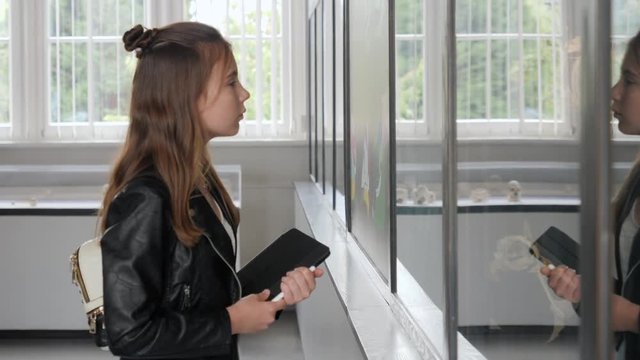 Teenager cute girl makes draw sketches in graphic tablet of animal skeletons in museum of nature as part of school task of biology