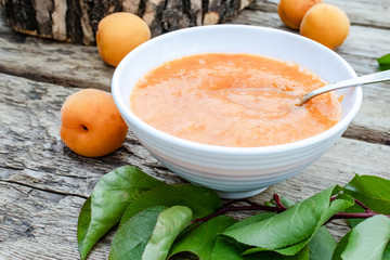 Apricot jam in a white bowl next to ripe apricot and green leaves on a wooden surface.