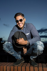 Young man portrait outdoors during twilight. He smiles, crouches and looks to the camera, has a hat in his hands and is wearing sunglasses