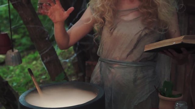 charming sorceress with blond hair and flower wreath on head preparing a healing decoction for lady, woman wearing old gray rag dress with book in her hands, heathen woman waving hand over cauldron