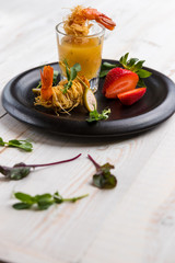 Crispy Shrimp in Kataifi Crust and Thyme with Champagne Sauce in glasses and strawberries lie on a black plate on a white woody background