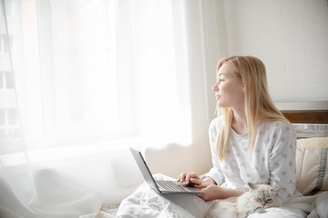 Fototapeta premium Blonde Giirl typing and using laptop hygge with fluffy cat, pajama and blanket. Hugge and lagom concept comfort and simple life . Horizontal copyspace