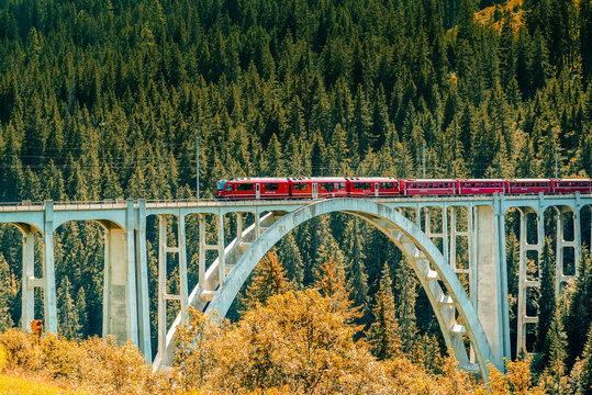 red train crosses a long viaduct across a deep ravine in the Swiss Alps
