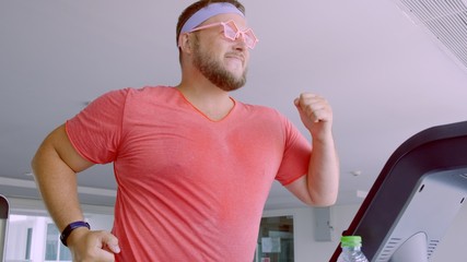 Playful fat man in a pink T-shirt and pink glasses is engaged in fitness in the gym.