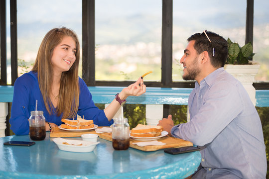 Couple during lunch at a park outdoors. Both are latinamericans in their twenties. They are wearing casual clothes, are eating sandwiches and drinking pop soda. The lady offers him some french fries. - Powered by Adobe