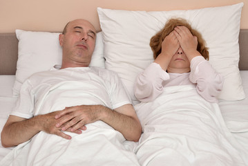 Mature couple in bed. Husband sleeping while his unhappy and depressed wife is covering her eyes with hands. Family crisis concept.
