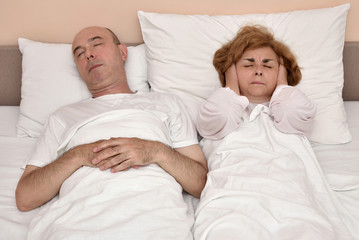 Couple in bed. Man snoring while woman is covering her ears with hands.