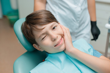 Patient in dental chair. Little boy having dental treatment at dentist's office. Child touch his cheek suffering from painful toothache.