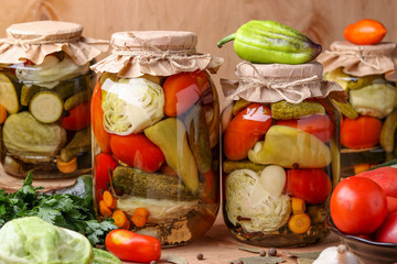 Assorted pickled vegetables in jars: cucumbers, tomatoes, cabbage, zucchini and peppers with garlic, dill and bay leaves in jars on a wooden background, horizontal photo