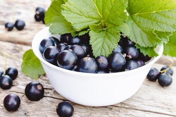 Fresh ripe currant berries in a white bowl on wooden background near green leaves. Juicy fruits currant. Black currant