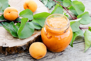Apricot jam in a glass jar next to a ripe apricot and green leaves on a wooden surface.