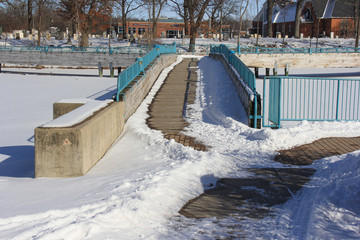 Obraz premium Snow & ice covered bridge with turquoise fencing, crossing a snow covered frozen lake. Trees and brick buildings in the backgroudn.